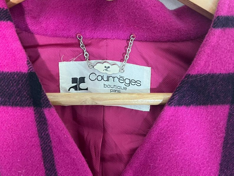 May include: Close-up of a magenta and black plaid coat. The coat features a deep V-neck and a label that reads "Courr&egrave;ges boutique paris". The coat is hanging on a wooden hanger.