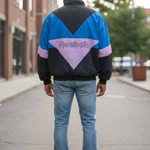 May include: A black, blue, and lavender Reebok windbreaker jacket. The jacket has a color block design with the Reebok logo across the back. The person is wearing light blue jeans and white sneakers.