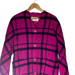 May include: A magenta and black plaid coat with a V-neck and three decorative buttons. The coat has a classic design with a tailored fit and two front pockets. The coat is hanging on a wooden hanger.