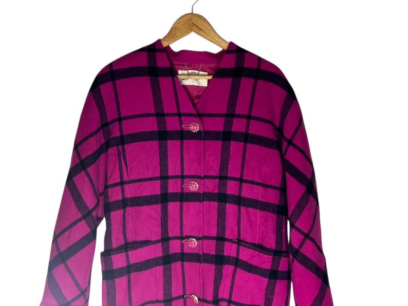 May include: A magenta and black plaid coat with a V-neck and three decorative buttons. The coat has a classic design with a tailored fit and two front pockets. The coat is hanging on a wooden hanger.