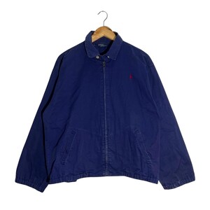 May include: A navy blue jacket with a full zipper closure and a small red logo on the chest. The jacket has a collared neckline and two front pockets. The jacket is hanging on a wooden hanger.