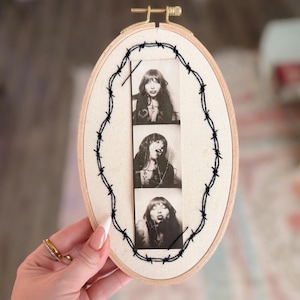 May include: An oval embroidery hoop with a black and white photo strip of a woman. The photo strip is surrounded by black barbed wire embroidery. The hoop is held by a hand with a gold ring.