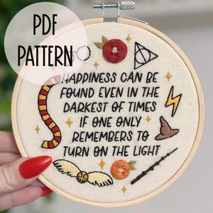 May include: A white embroidery hoop with a beige fabric background featuring a quote: "Happiness can be found even in the darkest of times if one only remembers to turn on the light". The quote is surrounded by various embroidered images including a lightning bolt, a witch's hat, a wand, a golden snitch, and a Hogwarts house crest.