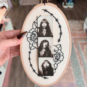 Könnte beinhalten: Ovaler Stickrahmen mit cremefarbenem Stoffhintergrund. Das Design zeigt einen Schwarz-Weiß-Fotostreifen einer Frau, umgeben von schwarzen gestickten Rosen und Stacheldraht.