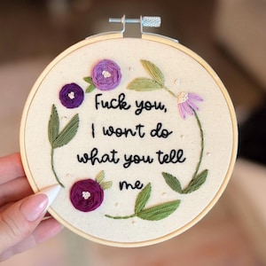 Puede incluir: Arte de aro bordado con el texto "Fuck you, I won't do what you tell me." El diseño presenta flores moradas y hojas verdes. El bordado está en un aro de madera.