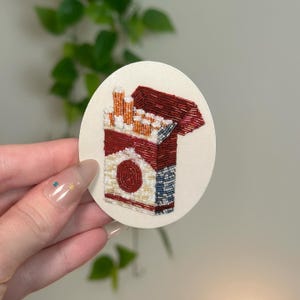 May include: Embroidered patch featuring a pack of cigarettes. The patch is round with a cream-colored background. The cigarette pack is red and white with a detailed design. The cigarettes are visible at the top.