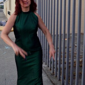 May include: A woman with long red hair is wearing a green halter dress and standing in front of a metal fence.