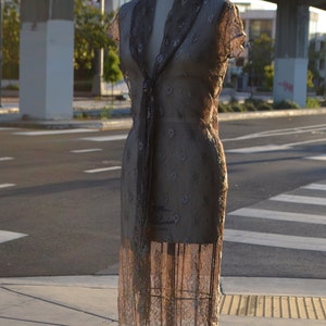 May include: A brown lace dress with a V-neckline and short sleeves is displayed on a mannequin. The dress has a delicate floral pattern and is displayed on a metal stand.
