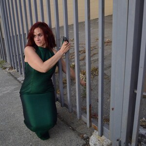 May include: A woman in a green dress stands in front of a metal fence. She is holding a pair of pliers in her right hand.