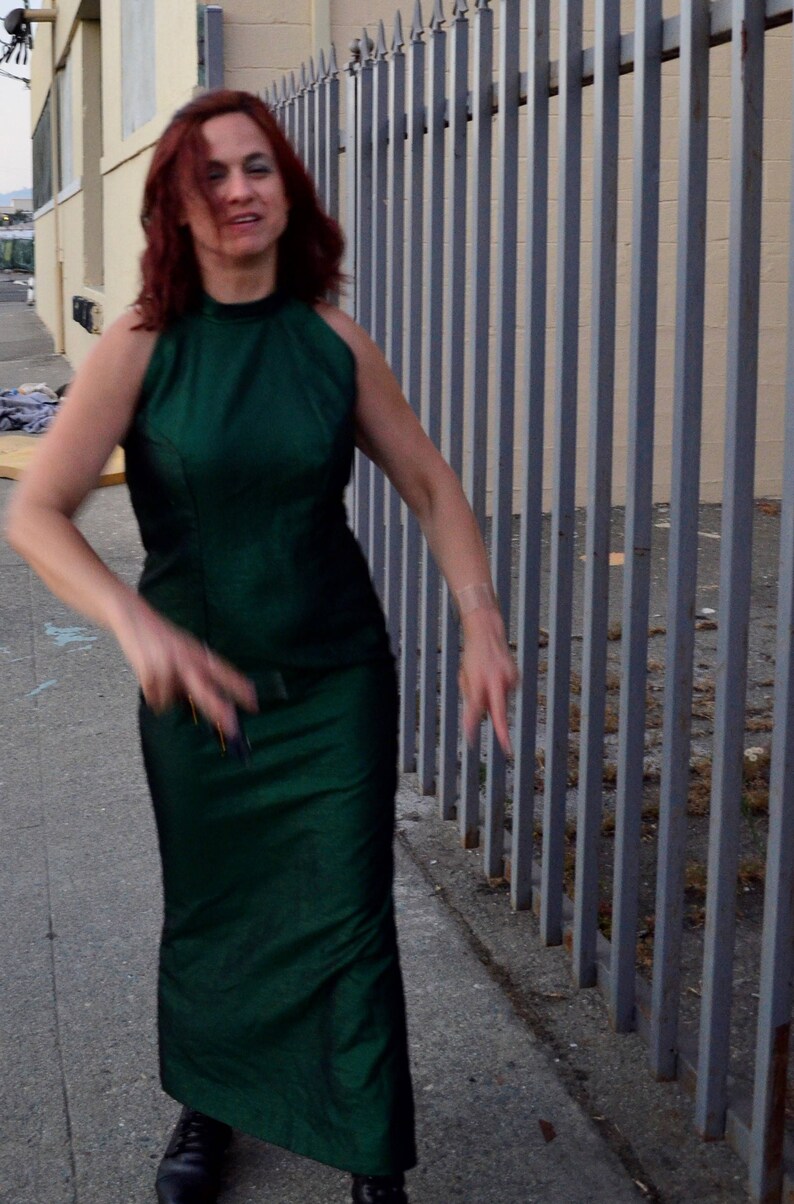 May include: A woman with long red hair is wearing a green halter dress and standing in front of a metal fence.