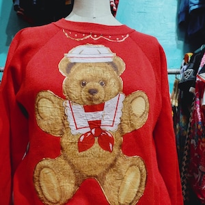 May include: Red sweatshirt with a brown teddy bear wearing a sailor hat and a red bow tie. The bear is surrounded by a white collar with a red stripe.