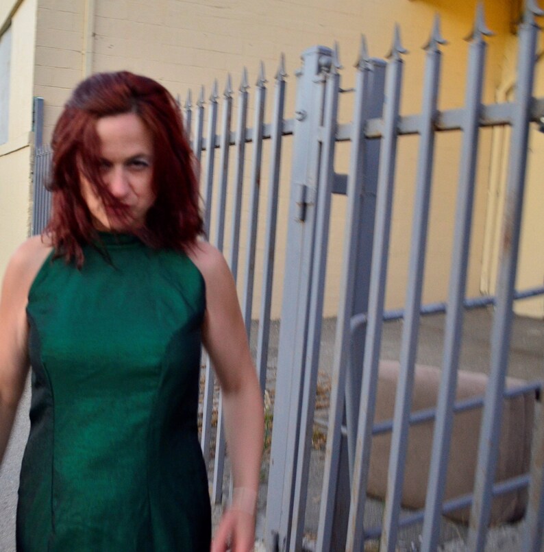 May include: A woman with red hair is wearing a green halter top dress. She is standing in front of a metal fence.