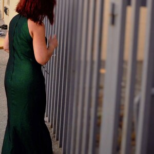 May include: A woman in a dark green dress stands in front of a metal fence. The fence is made of vertical bars and is painted a light gray color.
