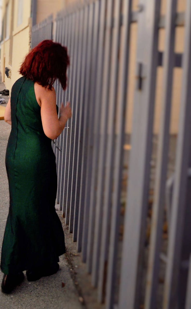 May include: A woman in a dark green dress stands in front of a metal fence. The fence is made of vertical bars and is painted a light gray color.