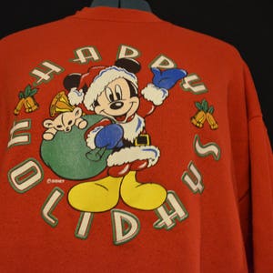 Könnte beinhalten: Roter Pullover mit einem Mickey-Mouse-Motiv. Mickey trägt einen Weihnachtsmann-Hut und hält eine grüne Tasche. Der Text "Happy Holidays" ist um Mickey herum geschrieben.