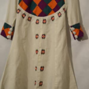 Puede incluir: Un vestido blanco con un corpiño a cuadros en tonos de morado, naranja y verde. El vestido tiene flores bordadas en la parte delantera y botones blancos en las mangas.