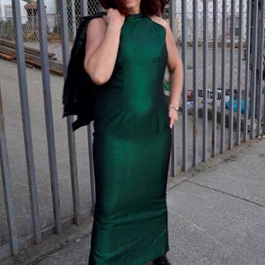 May include: A woman wearing a green halter-neck dress with a black leather jacket draped over her shoulder. She is standing in front of a metal fence.