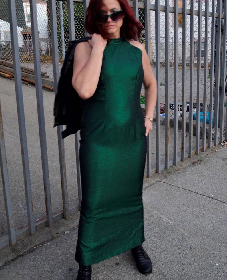 May include: A woman wearing a green halter-neck dress with a black leather jacket draped over her shoulder. She is standing in front of a metal fence.