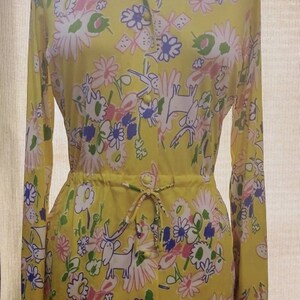 May include: A yellow long-sleeved shirt with a floral print featuring white, pink, blue, and green flowers and stylized animal figures. The shirt has a button-up front and a drawstring waist.