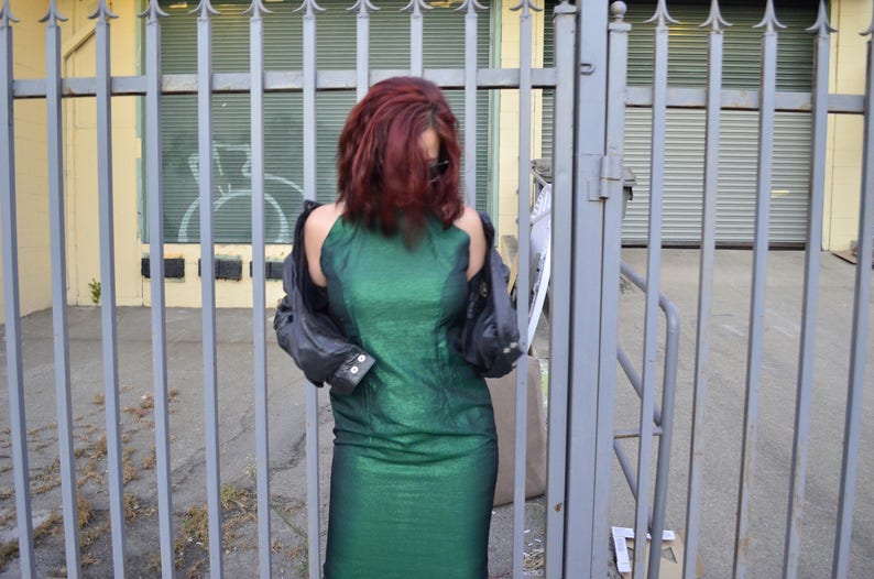 May include: A woman wearing a green sparkly dress and a black leather jacket stands in front of a metal fence.
