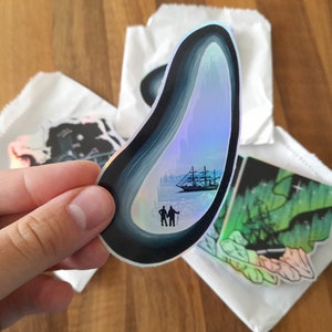 Puede incluir: Una pegatina holográfica con una silueta en blanco y negro de dos personas de pie en un paisaje nevado con un barco en el fondo.