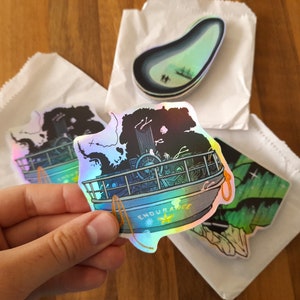 May include: A set of four iridescent holographic stickers featuring a ship, a crescent moon, and a green aurora borealis. The stickers are packaged in white paper envelopes.