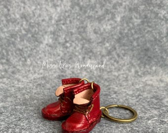 Puede incluir: Un par de botas en miniatura de cuero rojo unidas a un llavero dorado. Las botas tienen cordones y una solapa doblada en la parte superior.