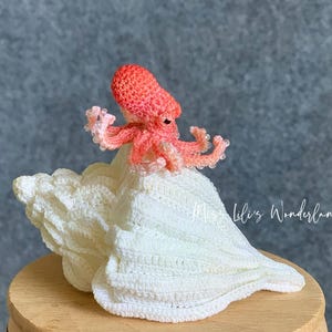 May include: A handmade crochet octopus in coral pink sits atop a white crochet seashell. The octopus has eight tentacles and is perched on a large, textured shell. The background is a muted gray. The text "Lili's Wonderland" is in the bottom right corner.