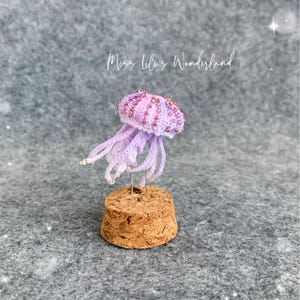 May include: A miniature crocheted jellyfish in light purple with pink beaded accents on the bell, featuring dangling tentacles. The jellyfish is mounted on a cylindrical cork base. The text Miss Lili's Wonderland is visible above the object.