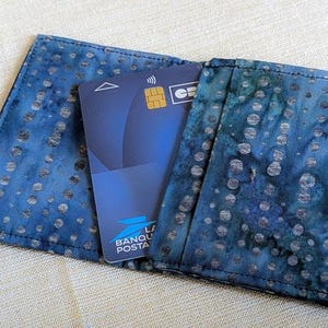 Może przedstawiać: Niebieski materiałowy portfel z wzorem w stylu batik, trzymający niebieską kartę kredytową. Portfel jest otwarty, pokazując kartę z napisem "BANQUE POSTALE". Tkanina ma wzór w kropki.