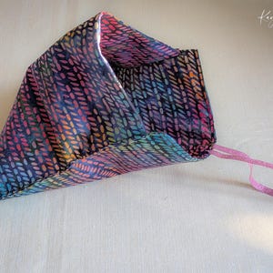 Pode incluir: Uma bolsa de tecido colorida com um padrão repetitivo de pequenas formas ovais em tons de azul, rosa, roxo e verde. A bolsa tem uma fita rosa presa. O tecido é dobrado para criar uma forma triangular.