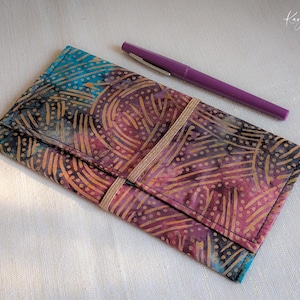 Op de afbeelding: Een batikstoffen portemonnee en een paarse pen. De portemonnee heeft een kleurrijk patroon in blauw, paars en goud, vastgezet met een gouden elastiek. De pen is effen paars van kleur.
