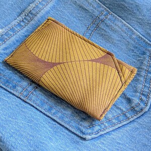 Könnte beinhalten: Ein senfgelbes und braun gemustertes Portemonnaie liegt in der Gesäßtasche einer hellblauen Jeans. Das Portemonnaie hat ein geometrisches Design mit radialen Linien und einem geschwungenen Mittelmuster.