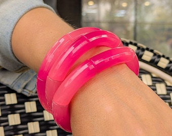 Può includere: Tre braccialetti rosa vibranti impilati su un polso. I braccialetti hanno una finitura liscia e lucida e un effetto sfumato, che passa da un rosa più chiaro a una tonalità più profonda e satura. I gioielli sono un'audace dichiarazione di moda.