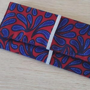 Op de afbeelding: Een rechthoekige clutch portemonnee met een rode achtergrond en een blauw-wit bloemenpatroon. Een zilveren elastische band loopt om het midden. De portemonnee is opgevouwen en lijkt van stof te zijn.