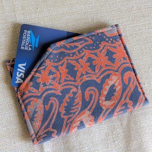 May include: A fabric card holder with a blue and orange floral pattern. A blue credit card with the word "VISA" is partially visible in the pocket. The card holder is on a neutral-coloured surface.
