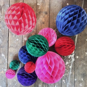 May include: A collection of colorful honeycomb paper balls in various sizes and shades, including red, blue, green, pink, and purple. The decorative spheres are arranged on a rustic wooden surface, creating a vibrant and festive display.