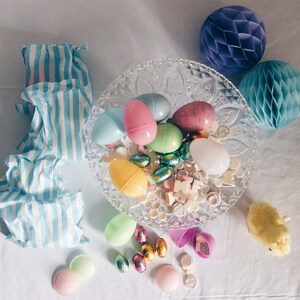 Pu&ograve; includere: Una ciotola di vetro trasparente piena di uova di Pasqua di plastica colorate, caramelle e piccoli oggetti decorativi. La ciotola &egrave; circondata da carta velina a strisce blu e bianche, una palla a nido d'ape viola, un giocattolo di pulcino bianco e altre decorazioni pasquali.