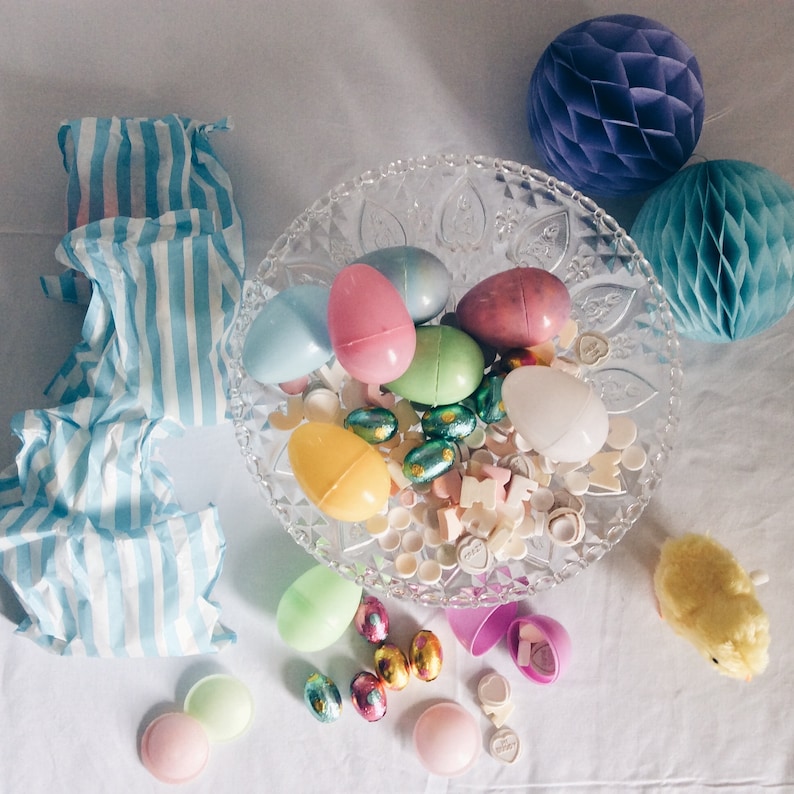 Pu&ograve; includere: Una ciotola di vetro trasparente piena di uova di Pasqua di plastica colorate, caramelle e piccoli oggetti decorativi. La ciotola &egrave; circondata da carta velina a strisce blu e bianche, una palla a nido d'ape viola, un giocattolo di pulcino bianco e altre decorazioni pasquali.