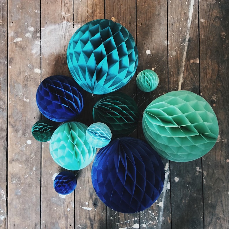 May include: An assortment of decorative paper honeycomb balls in various shades of blue and green. The balls are arranged on a rustic wooden surface, creating a visually appealing display. The sizes of the balls vary, adding depth to the composition.