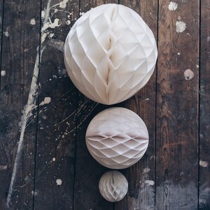 May include: Three paper honeycomb balls in varying sizes, arranged on a weathered wooden surface. The largest ball is at the top, with the other two below. The balls are a soft, off-white color, and the wood has paint splatters.