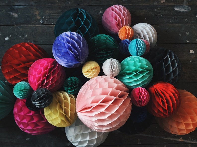 May include: A collection of colorful, honeycomb paper balls in various sizes and hues, including red, pink, blue, green, and yellow. These decorative spheres are arranged on a dark wooden surface, creating a vibrant and textured display.