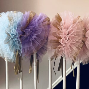 Op de afbeelding: Drie decoratieve tule pom-poms in pastelkleuren: lichtblauw, lavendel en roze. Elke pom-pom heeft een gouden mesh accent en gouden lint streamers. De pom-poms worden op een witte balustrade getoond.