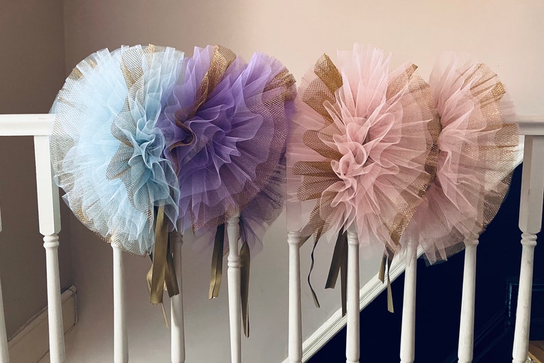 Op de afbeelding: Drie decoratieve tule pom-poms in pastelkleuren: lichtblauw, lavendel en roze. Elke pom-pom heeft een gouden mesh accent en gouden lint streamers. De pom-poms worden op een witte balustrade getoond.