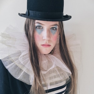 Op de afbeelding: Een persoon draagt een zwarte bowlerhoed, een witte en gouden gefranjerde kraag en een zwart-wit gestreept shirt. De persoon heeft blauwe oogmake-up en roze blush op de wangen, wat een clownachtig uiterlijk cre&euml;ert.