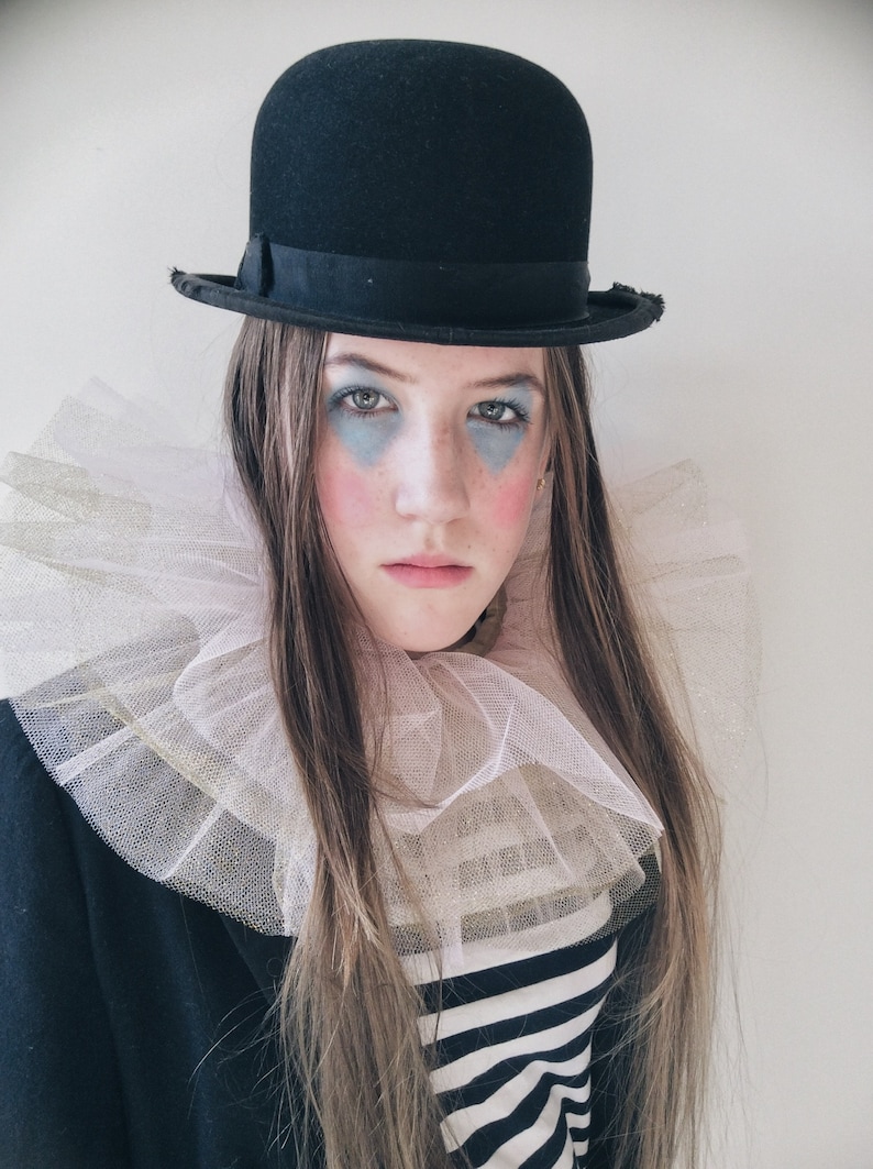 Op de afbeelding: Een persoon draagt een zwarte bowlerhoed, een witte en gouden gefranjerde kraag en een zwart-wit gestreept shirt. De persoon heeft blauwe oogmake-up en roze blush op de wangen, wat een clownachtig uiterlijk cre&euml;ert.