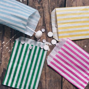 Pu&ograve; includere: Quattro sacchetti di carta a righe in diversi colori, tra cui blu, giallo, verde e rosa. I sacchetti sono disposti su una superficie di legno con spolverata di caramelle bianche sparse intorno.
