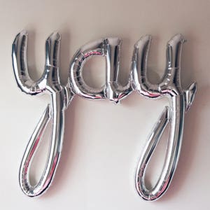 Puede incluir: Globos de aluminio plateado que deletrean la palabra "yay" en una fuente cursiva.