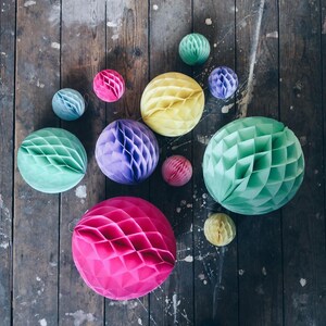 May include: A collection of colorful honeycomb paper balls in various sizes and shades, including pink, yellow, green, blue, and purple. The decorative spheres are arranged on a weathered wooden surface, creating a vibrant and playful display.