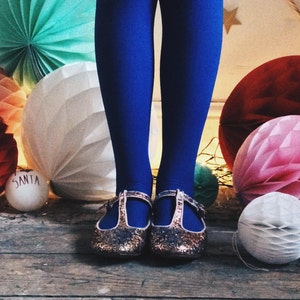 May include: A festive scene featuring blue tights and glittery gold shoes with T-straps. The background includes colorful paper honeycomb decorations in red, pink, white, and teal, with a small ornament that says "SANTA".